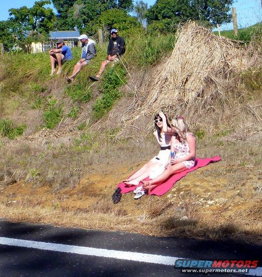 Four people sit on a grassy hill overlooking a road; two women sit on a pink blanket in the foreground