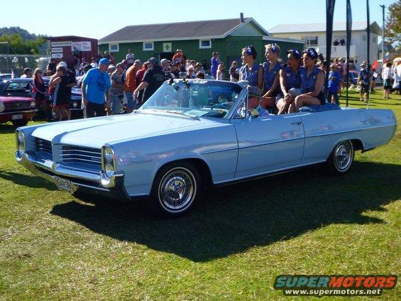Light blue 1965 Pontiac Catalina convertible with four women in blue outfits in the passenger seats. Crowd in background