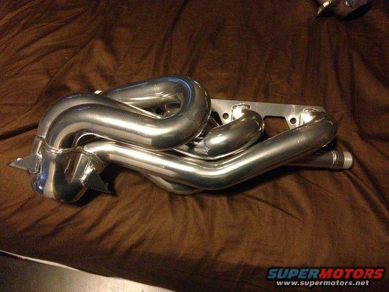 Shiny metal exhaust headers on a brown cloth surface. Text at bottom reads SUPERMOTORS