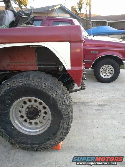 Big off-road tires on a red and white pickup truck, another red pickup behind it