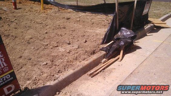 construction site with dirt, black plastic sheeting, and lumber near a curb. Supermotors.net logo in lower right