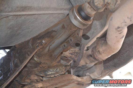 Underside of a rusty vehicle showing a driveshaft, exhaust pipe, and frame components