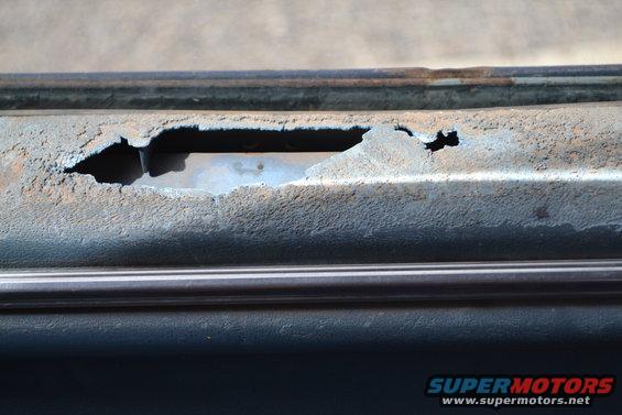 Close-up of heavily rusted metal panel with a large hole showing blue interior, on a dark blue vehicle