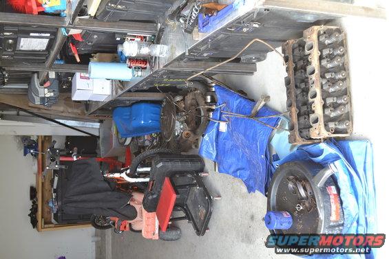 Engine parts including a cylinder head and oil pan on a garage floor with tarps and tools