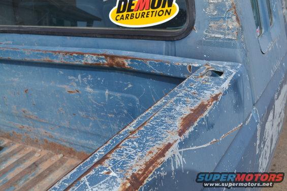 Blue pickup truck bed with rust and scratches. Sticker on rear window says 'DEMON CARBURETION'