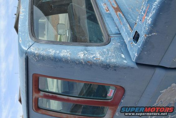 Blue truck cab with peeling paint and rust. Side view shows a small rectangular mirror inside and a red-trimmed window
