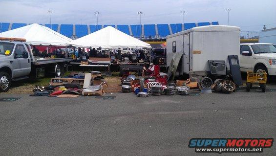 Car parts including wheels, doors, and trim are displayed on the ground next to a white trailer and a white pickup truck