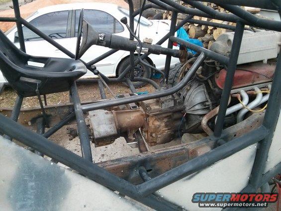 Custom buggy frame with engine, transmission, and steering column. White car in background