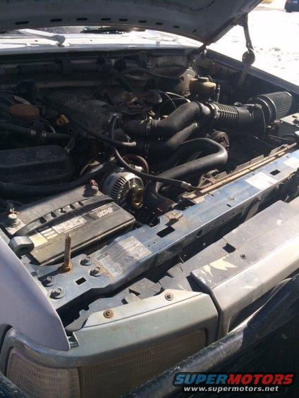 Engine bay of a light-colored SUV with the hood open. Visible components include the battery, alternator, and air intake