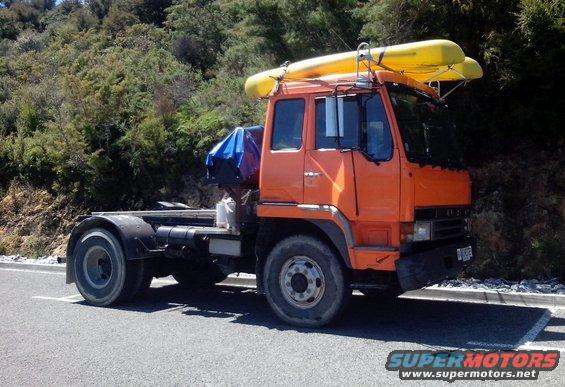Orange truck with two yellow kayaks on roof rack, parked on roadside with trees in background