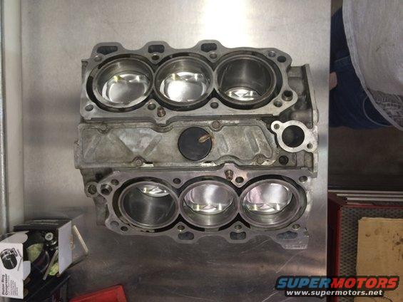 Engine block with six cylinders and pistons visible, sitting on a metal surface. A box labeled 'Piston Ring Compressor' is to the left