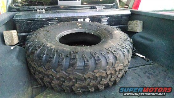 Spare off-road tire with aggressive tread, lying flat in the bed of a pickup truck next to a toolbox
