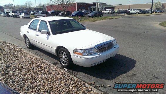 White sedan parked on a street, angled from the front-left. The car has chrome grille and alloy wheels