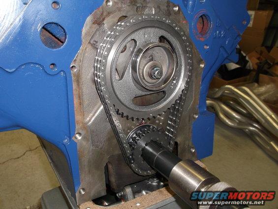 Close-up of engine timing chain and gears with blue block and exhaust headers in background. Supermotors.net logo visible