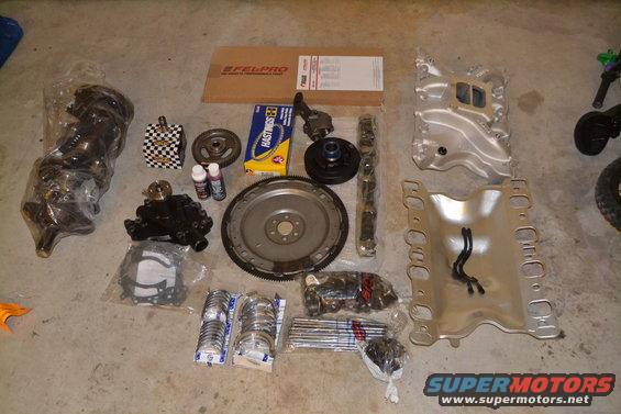Engine parts including crankshaft, intake manifold, exhaust manifolds, pistons, bearings, and gaskets laid out on concrete floor