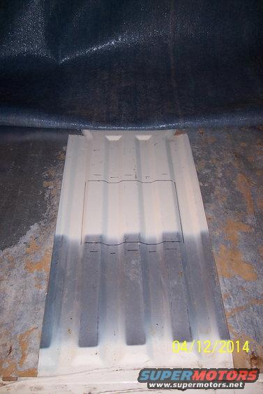 Corrugated metal floor panel with a rectangular cutout, possibly for access. A dark tarp is visible above