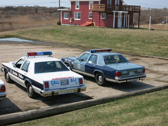 Two police cars, one white Boston Police and one blue State Police, parked outside a red building