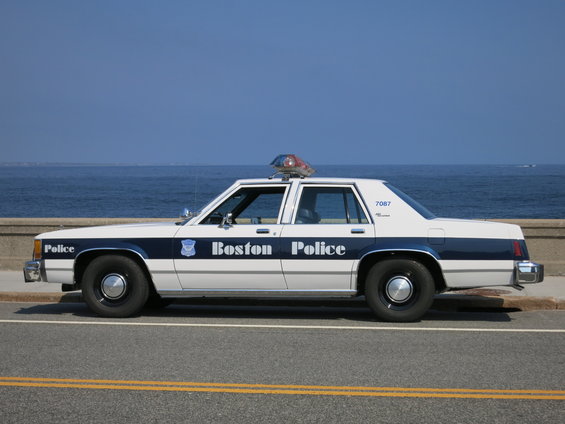 White 1991 Ford Crown Victoria police car with blue stripe and "Boston Police" text, parked by the ocean