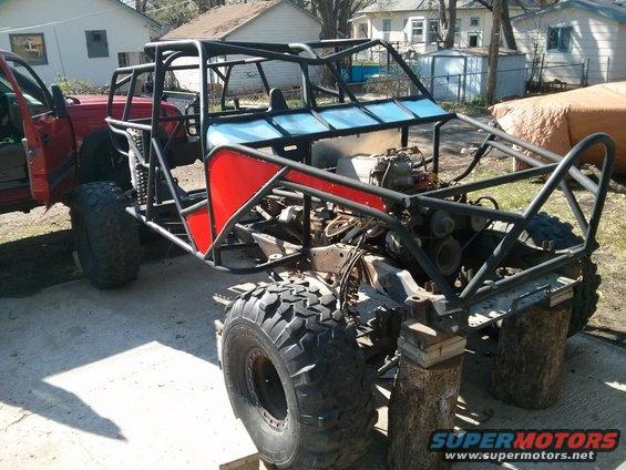 Custom buggy frame with red panels and large tires, sitting on blocks. Red truck cab visible on left