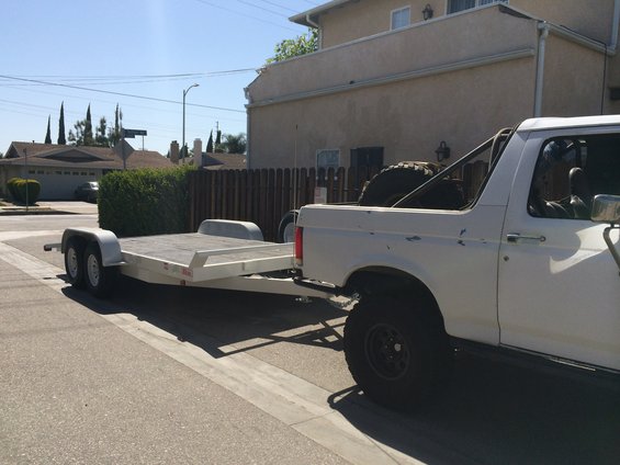 White pickup truck with a roll bar towing an empty flatbed trailer on a residential street