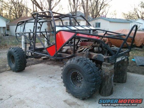 Custom buggy frame with red panels and large off-road tires, supported by wooden blocks