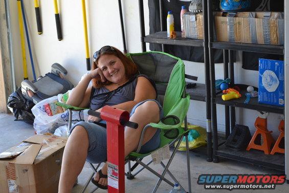 Woman in a green camping chair in a garage, with tools and shelves in the background. Supermotors logo visible