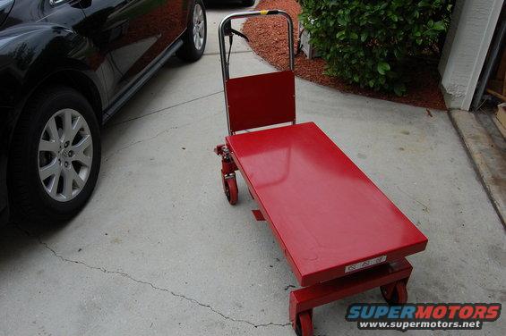 Red hydraulic lift table with wheels and a black car in the background