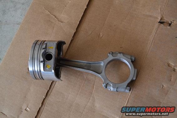 Piston and connecting rod assembly on a cardboard surface. Some yellow markings are visible on the piston
