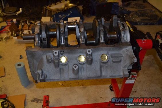 Engine block on a red stand, showing the crankshaft and connecting rods. Parts and tools are scattered in the background