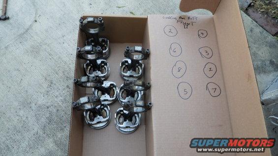 Cardboard box with eight pistons and connecting rods laid out, with handwritten numbers on the inside flap indicating cylinder order