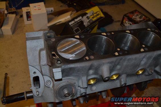 Engine block with cylinder bores visible, on a red stand. A "Supermotors.net" logo is in the lower right