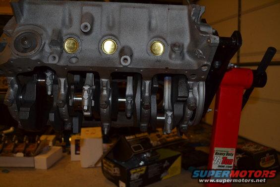 Engine block on a red engine stand, showing crankshaft and connecting rods. Gold plugs visible