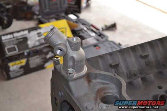 Engine block with a metal thermostat housing attached. Tools and a box labeled 'Intake Manifold' are in the background