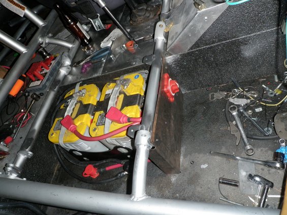 Dual yellow batteries with red terminals are mounted inside a vehicle with a metal roll cage. Tools and a beer bottle are also visible