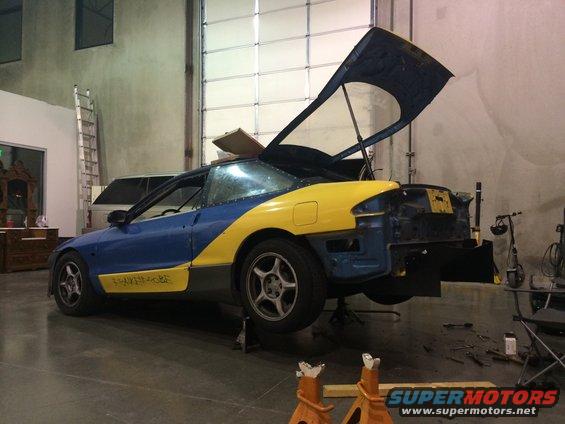 Blue and yellow car with hood open, on jack stands