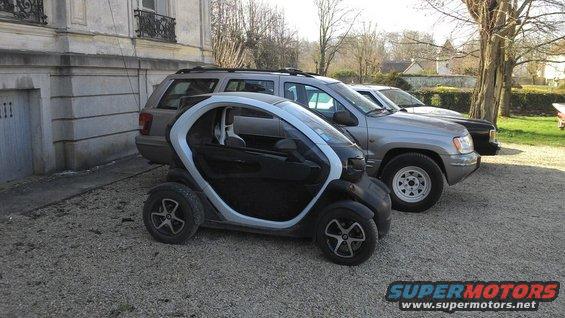 Small black and silver microcar parked in front of a silver SUV and a dark SUV