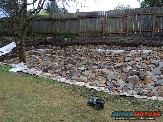Two toy trucks on grass and rocks near a wooden fence