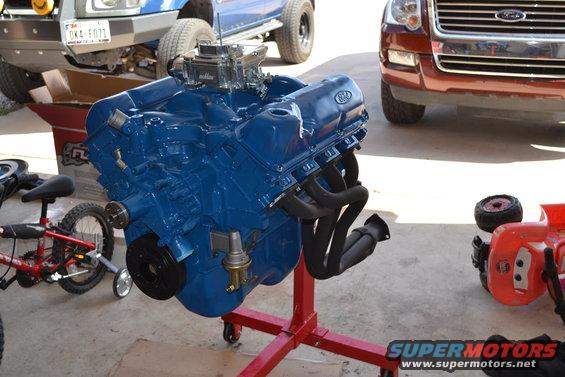 Blue V8 engine on a red stand, with parts of trucks and cars in background
