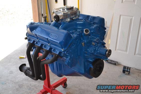 Blue Ford V8 engine on a red stand, with black exhaust headers and a chrome carburetor