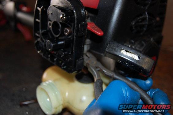 Close-up of blue-gloved hands using pliers to disconnect a fuel line from a black engine component with a yellow fuel tank