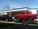 1978 Ford Bronco - A red 1978 Ford Bronco is loaded onto a trailer, towed by a black pickup truck in front of a building.