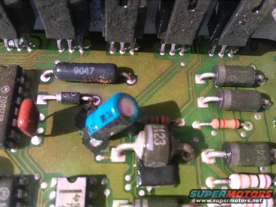 Close-up of a green circuit board with various electronic components including resistors, capacitors, diodes, and integrated circuits. Some components show signs of wear or corrosion