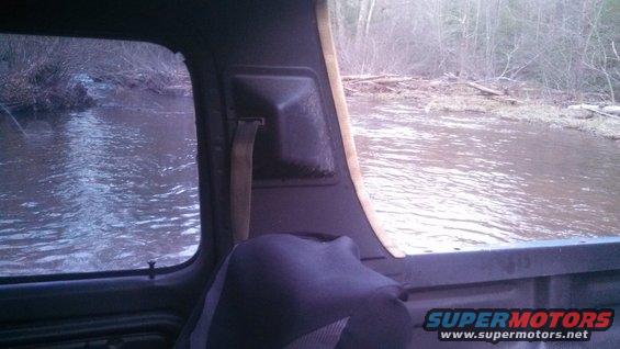 View from inside a vehicle, looking out the side window at a flooded area with trees and debris