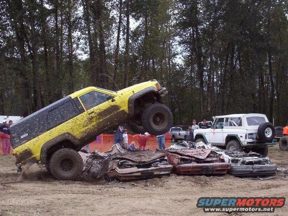 Yellow Jeep Cherokee is climbing over wrecked cars, with a white Ford Bronco in the background