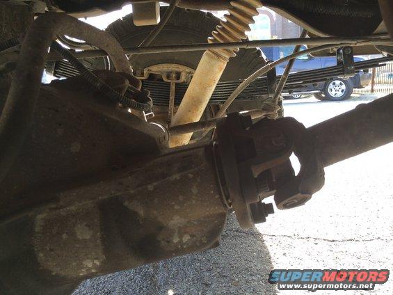 Underside view of a vehicle's rear axle, driveshaft, and leaf springs. Another truck is visible in the background