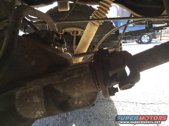 Close-up of a vehicle's rear axle, leaf springs, and shock absorber. A blue truck is visible in the background