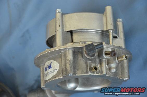 Close-up of a metal throttle body with a blue and white KM sticker. Part of a SuperMotors.net watermark is visible