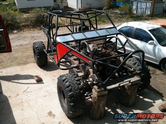 Off-road buggy chassis with roll cage, red side panels, and exposed engine, supported by logs. White sedan in background