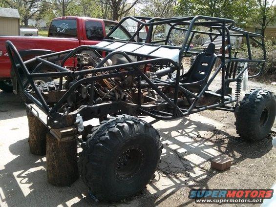 Custom buggy frame with large tires and exposed engine components, parked next to a red pickup truck