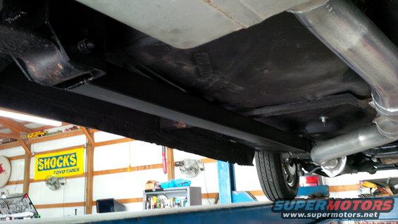 Underside view of a car showing the frame, exhaust pipes, and a rear wheel. A 'SHOCKS TOYO TIRES' sign is visible in the background